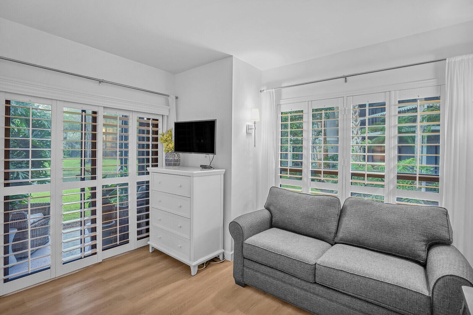 A living room features a gray sofa, white dresser with a television, and plantation shutters — 11863 Wimbledon Circle # 423, Wellington FL 33414, WELLINGTON, FL