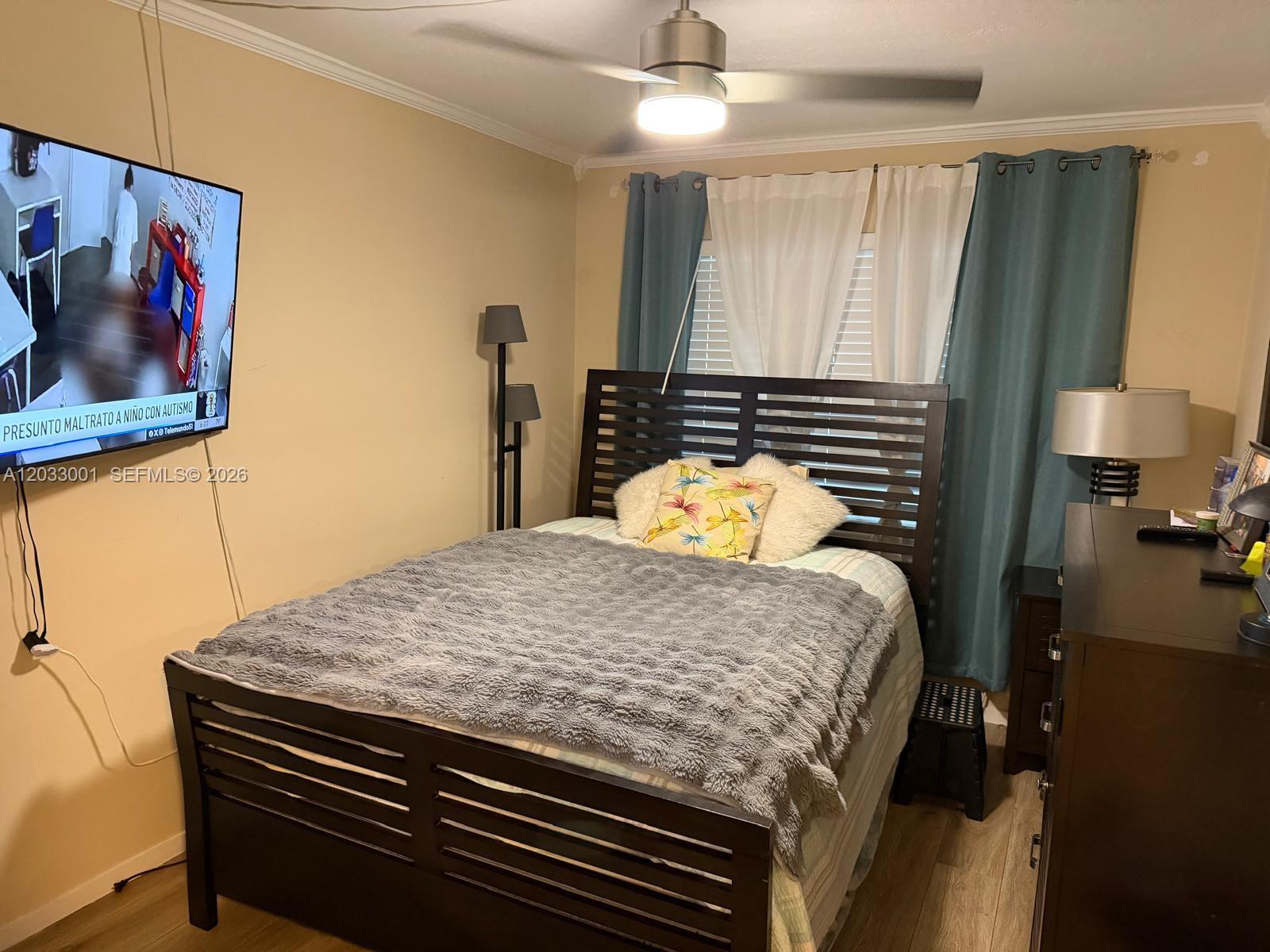 A bedroom features a dark wood bed frame, a wall-mounted television, and teal curtains — 500 Layne Blvd # 12, Hallandale Beach FL 33009, HALLANDALE BEACH, FL