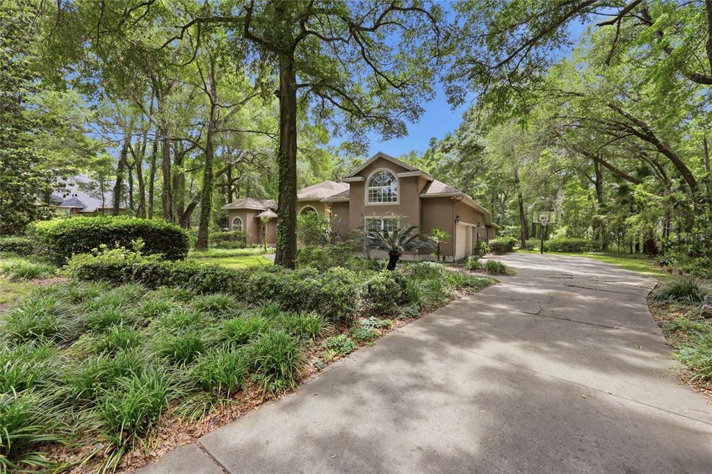 A tan two-story house with a concrete driveway sits among many mature trees — 4208 SW 96TH DRIVE, GAINESVILLE, FL