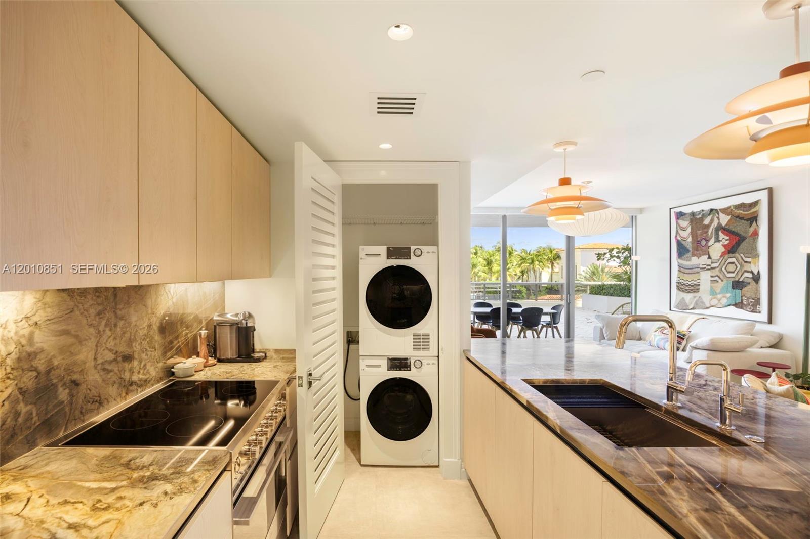 A modern kitchen features light wood cabinets, marble countertops, and a stacked washer-dryer unit — 5875 Collins Ave # 503, Miami Beach FL 33140, MIAMI BEACH, FL