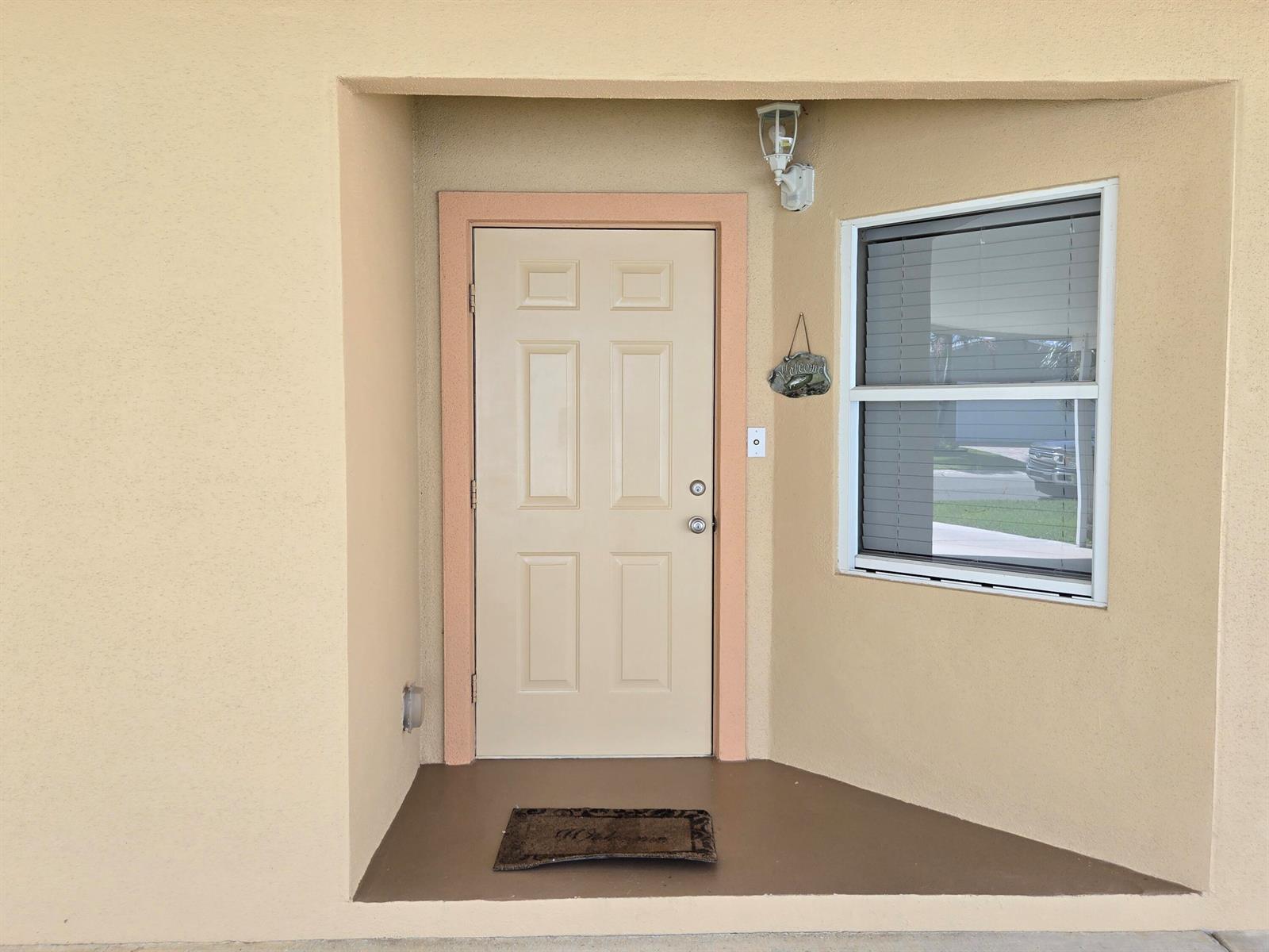 A beige front door with a window and a welcome mat sits under a light — 4311 SW 9th Way, Okeechobee FL 34974, OKEECHOBEE, FL