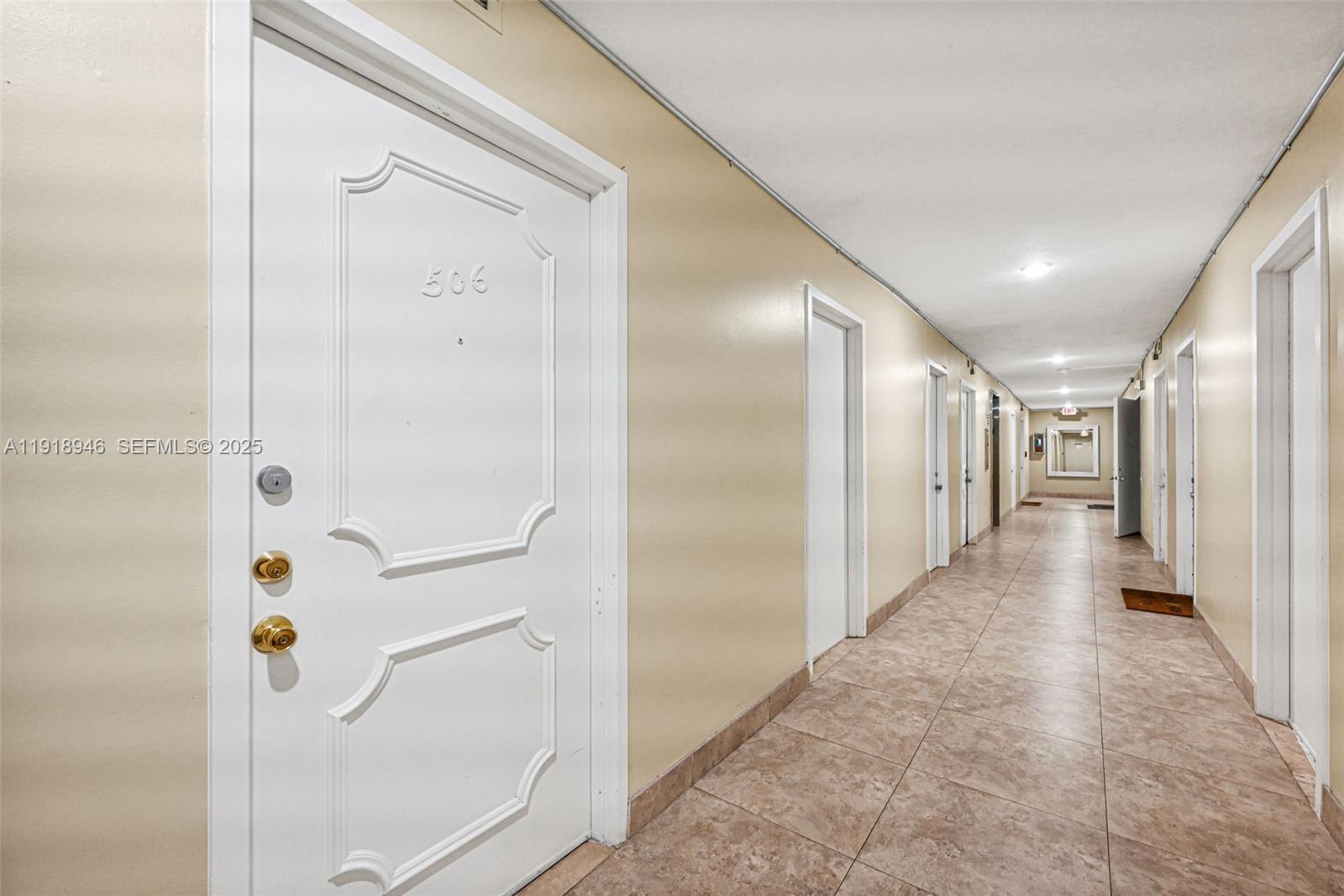 A long apartment hallway features beige walls, tiled floors, and several white doors — 1830 Dixieanna St # 506, Hollywood FL 33020, HOLLYWOOD, FL