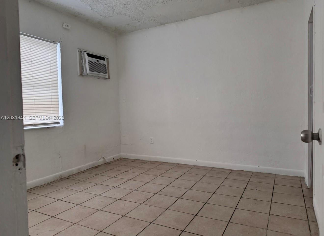 An empty room with white walls, tiled floors, a window with blinds, and a wall-mounted air conditioner — 1601 NW 62nd St # 26, Miami FL 33147, MIAMI, FL