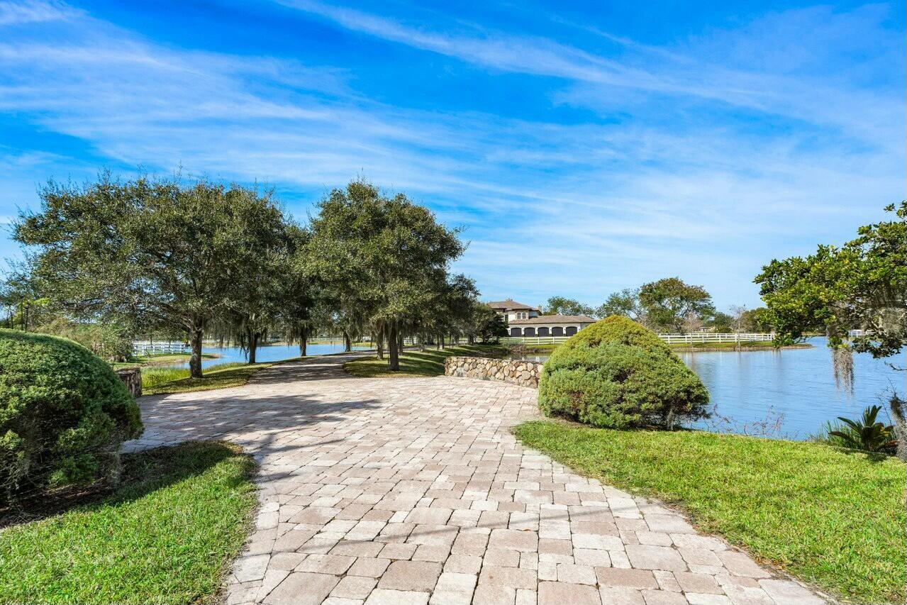 A paved walkway leads toward a house across a small lake surrounded by green trees — 10527 El Paraiso Place, Delray Beach FL 33446, DELRAY BEACH, FL