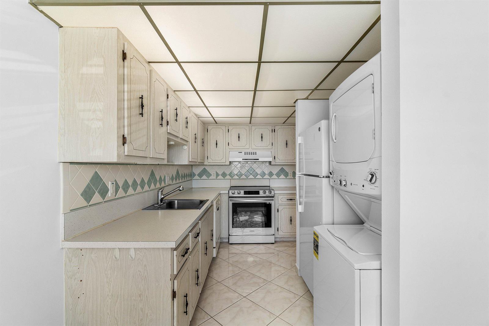 A galley kitchen features light wood cabinets, a tiled backsplash, and a stacked washer-dryer unit — 2835 SW 13th Street # 202, Delray Beach FL 33445, DELRAY BEACH, FL