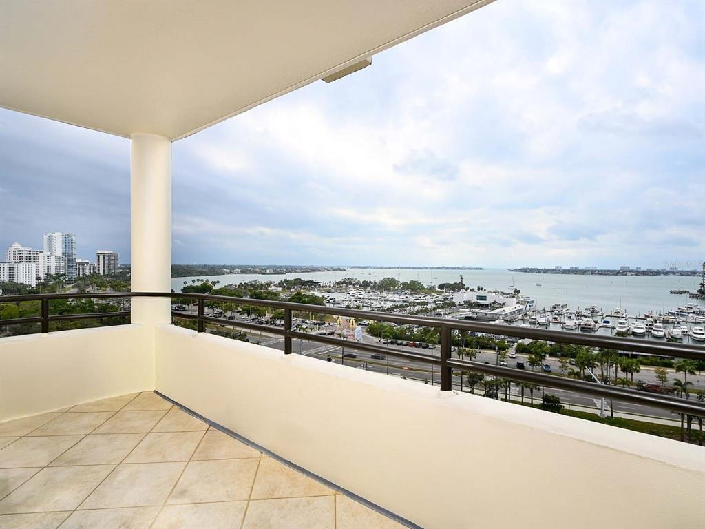 A balcony with tile flooring overlooks a marina, a coastal road, and a distant city skyline — 1255 N GULFSTREAM AVENUE UNIT 1106, SARASOTA, FL