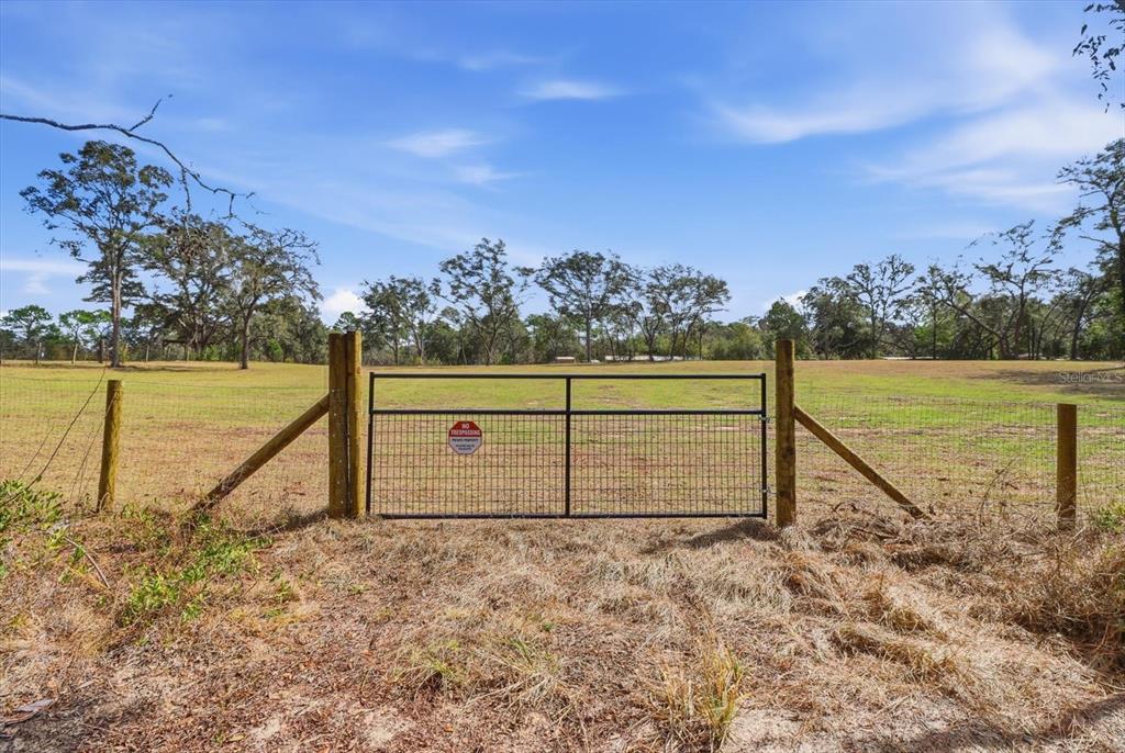 A metal gate stands in a wire fence bordering a large, grassy, tree-lined field — 2720 E FAWN COURT, INVERNESS, FL