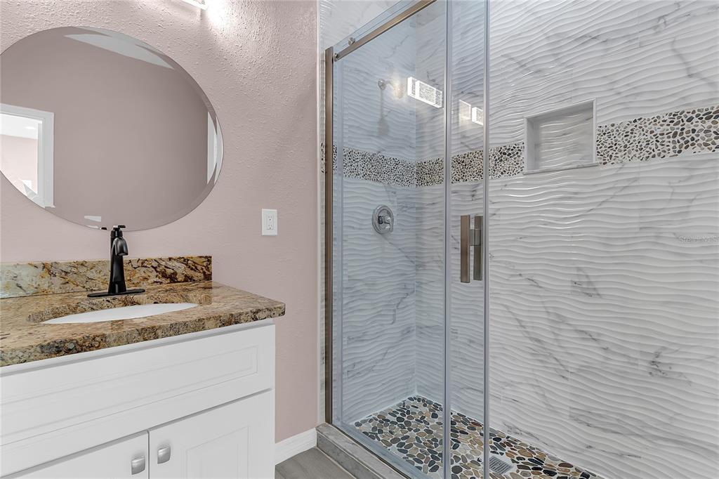 A bathroom features a granite vanity, a round mirror, and a glass-enclosed tiled shower — 707 HAMILTON STREET, NEW SMYRNA BEACH, FL