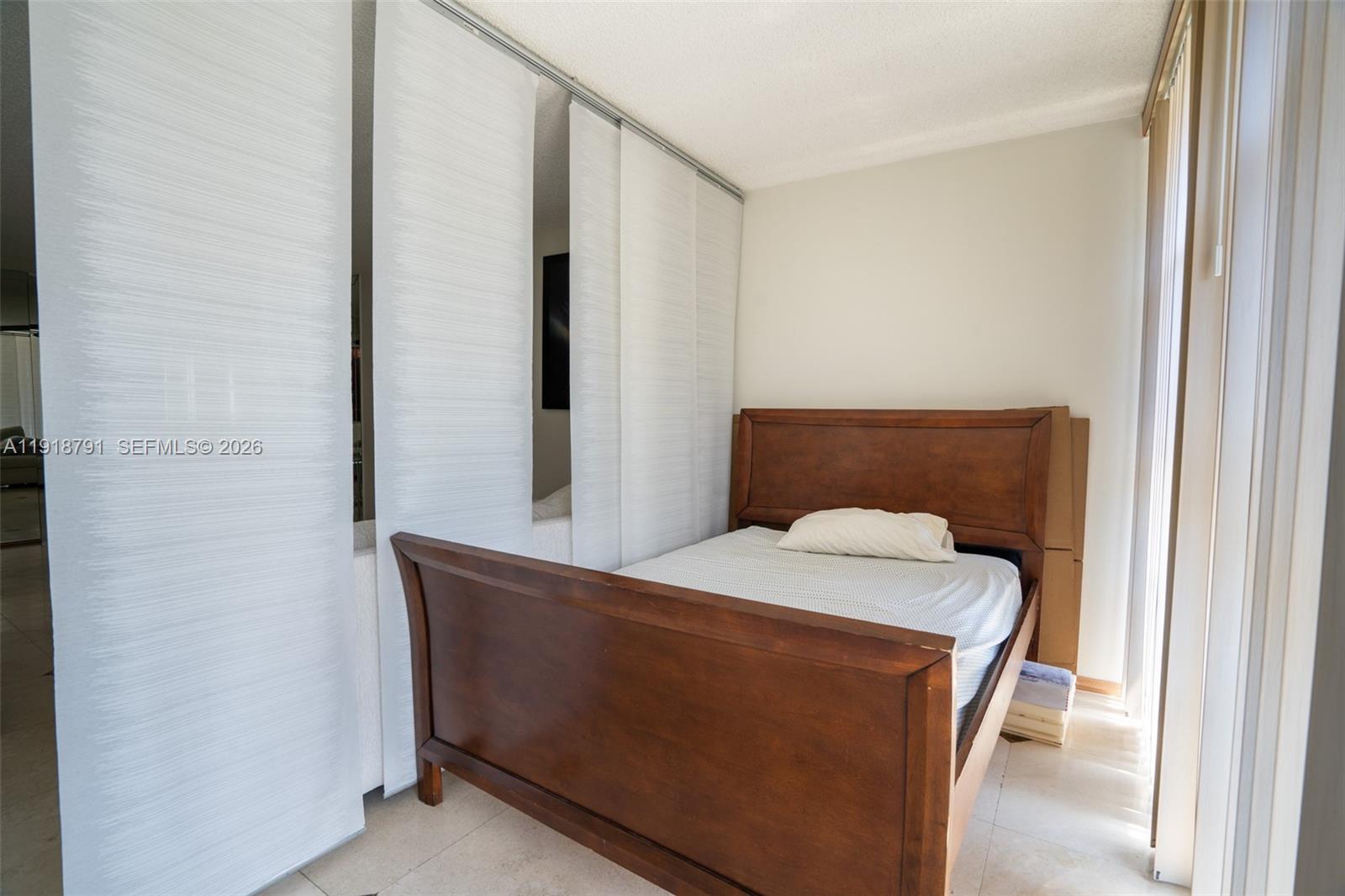 A bedroom features a wooden bed frame positioned against a wall with sliding panel blinds — 701 Three Islands Blvd # 506, Hallandale Beach FL 33009, HALLANDALE BEACH, FL