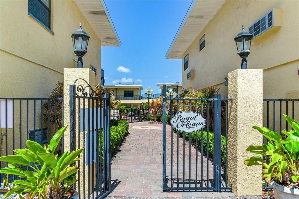 A gated brick walkway leads between two yellow apartment buildings toward a pool area — 16333 GULF BOULEVARD UNIT 113, REDINGTON BEACH, FL