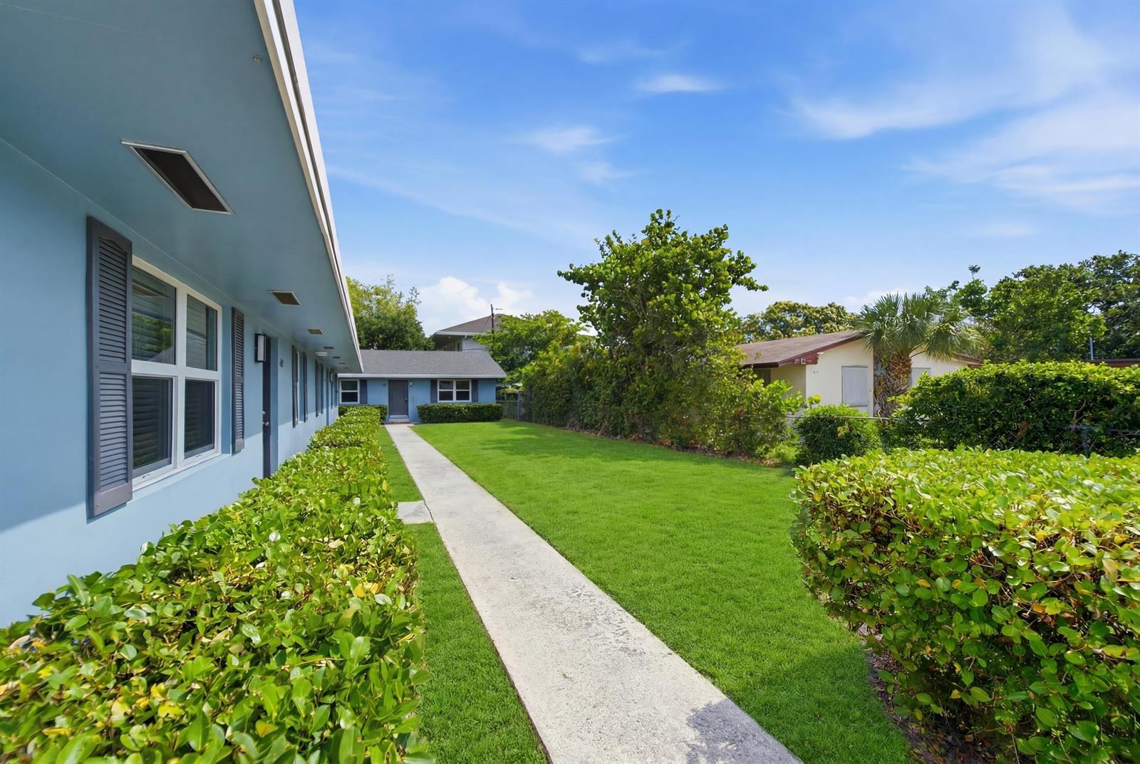 A concrete walkway leads between a blue building and a hedge toward a small house — 351 Wickline Boulevard, Lantana FL 33462, LANTANA, FL