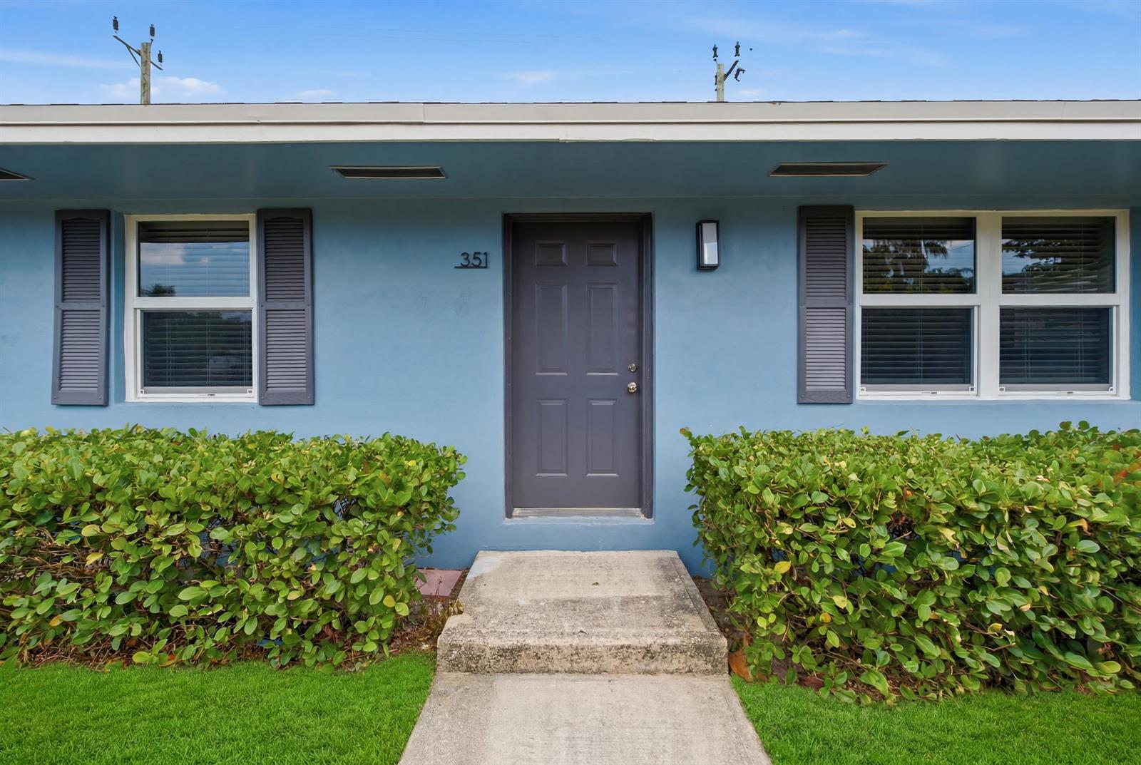 A blue exterior wall features a gray front door, two windows with shutters, and hedges — 351 Wickline Boulevard, Lantana FL 33462, LANTANA, FL