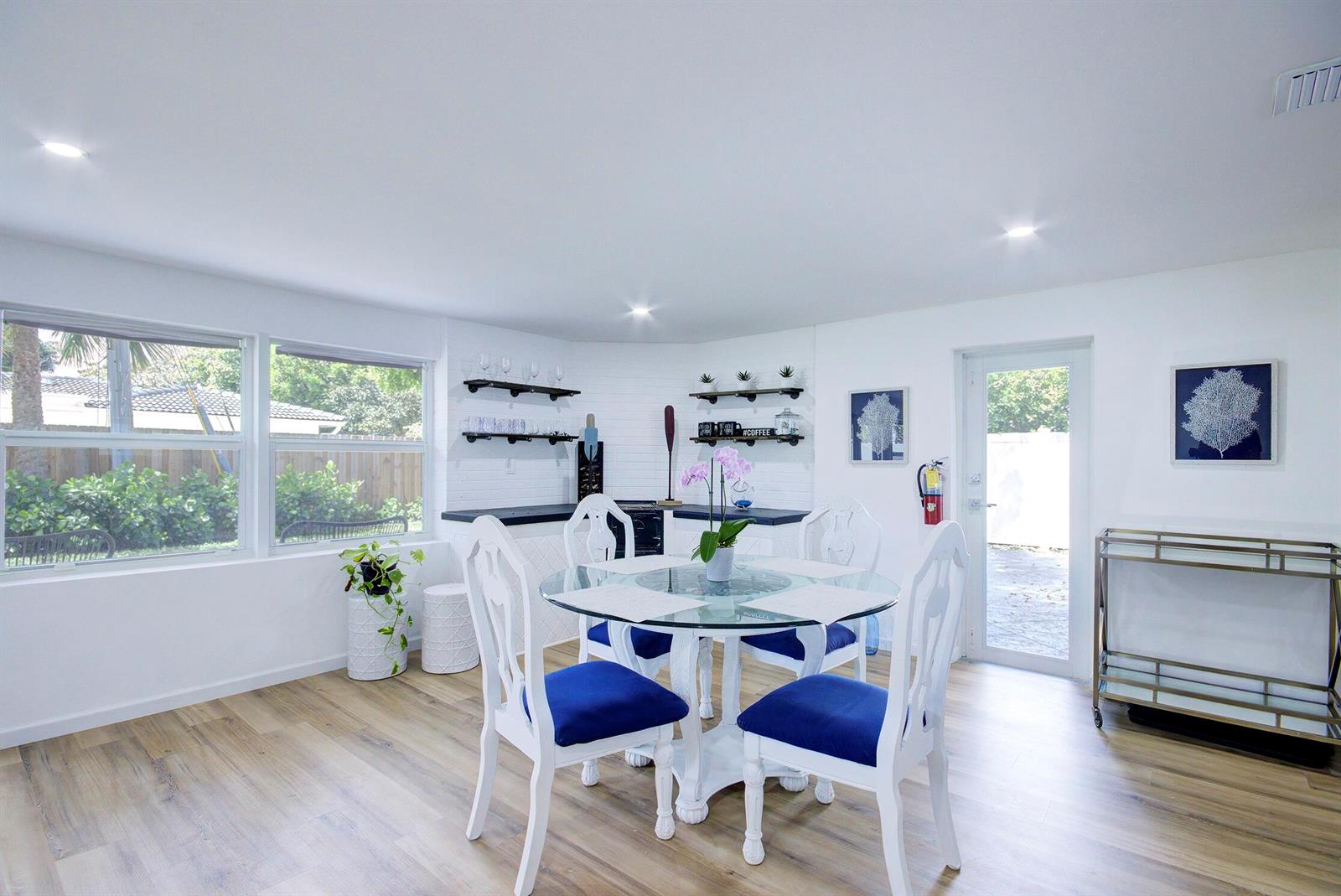 A dining area features a glass-topped table, blue-cushioned chairs, white brick walls, and wood-look flooring — 1950 NE 30th Street, Lighthouse Point FL 33064, LIGHTHOUSE POINT, FL