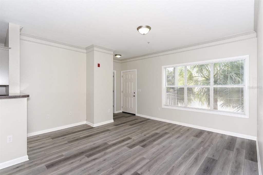 An empty living room features gray wood-look flooring, light walls, crown molding, and a large window — 4802 51ST STREET W UNIT 1009, BRADENTON, FL