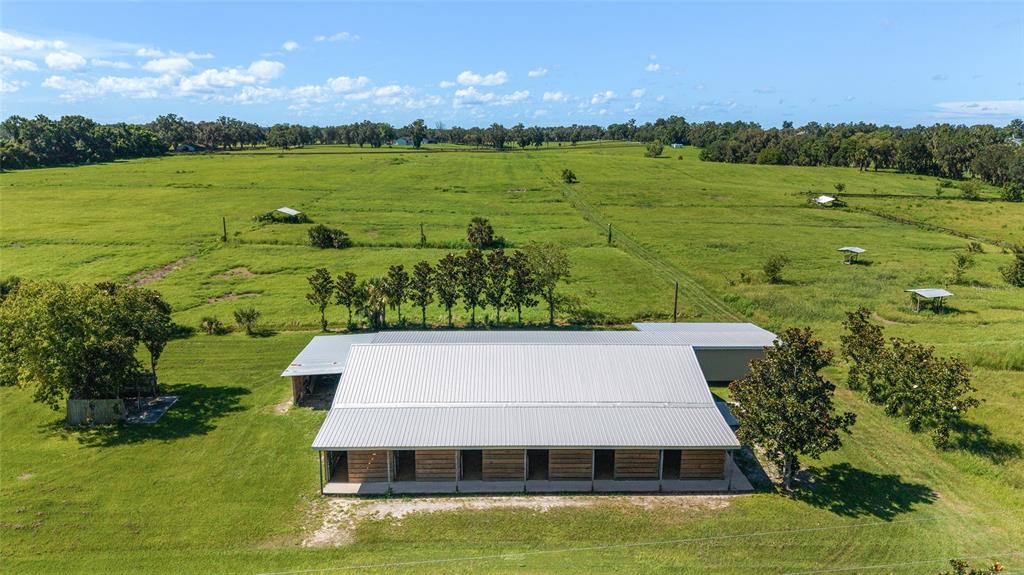 An aerial view shows a long, metal-roofed barn with multiple stalls situated in a large pasture — 5800 SE 145TH STREET, SUMMERFIELD, FL