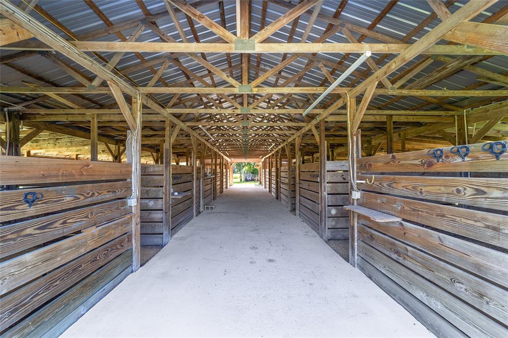 An open-air horse stable features a concrete aisle flanked by wooden stalls under a trussed roof — 5800 SE 145TH STREET, SUMMERFIELD, FL