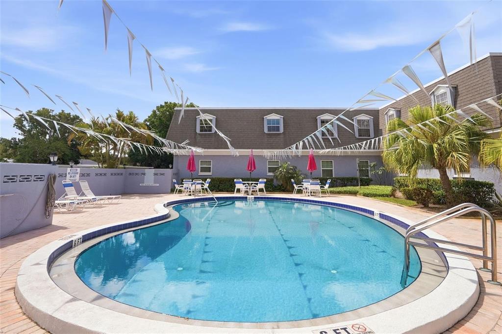 A kidney-shaped swimming pool sits in front of a two-story apartment building with patio furniture — 1717 BELLEAIR FOREST DRIVE UNIT A, CLEARWATER, FL
