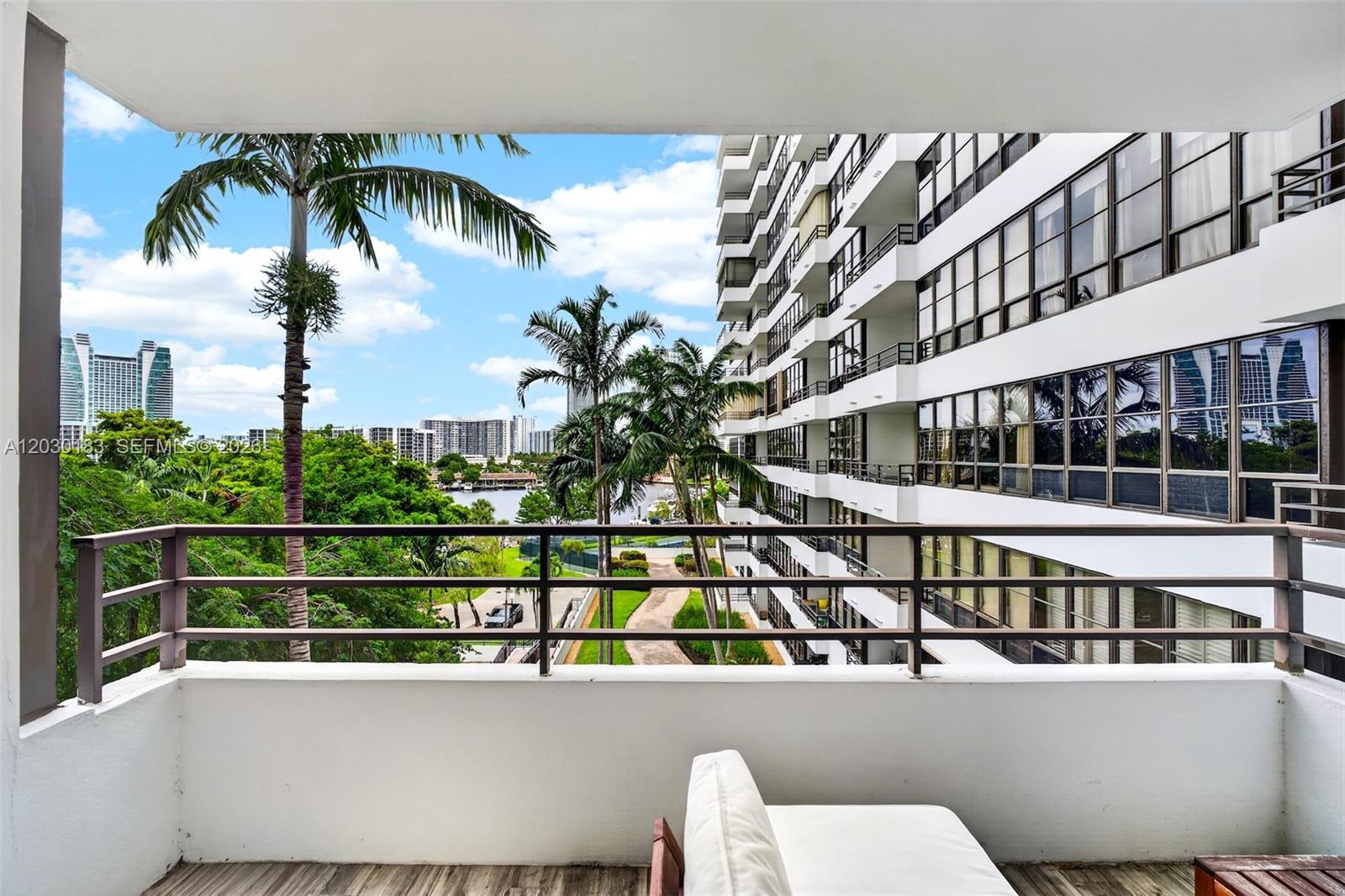 A balcony view features a white building, palm trees, and a distant cityscape — 500 Three Islands Blvd # 201, Hallandale Beach FL 33009, HALLANDALE BEACH, FL