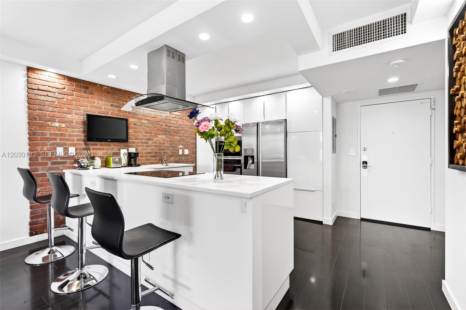 A modern kitchen features a white island, black bar stools, and an exposed brick wall — 500 Three Islands Blvd # 201, Hallandale Beach FL 33009, HALLANDALE BEACH, FL