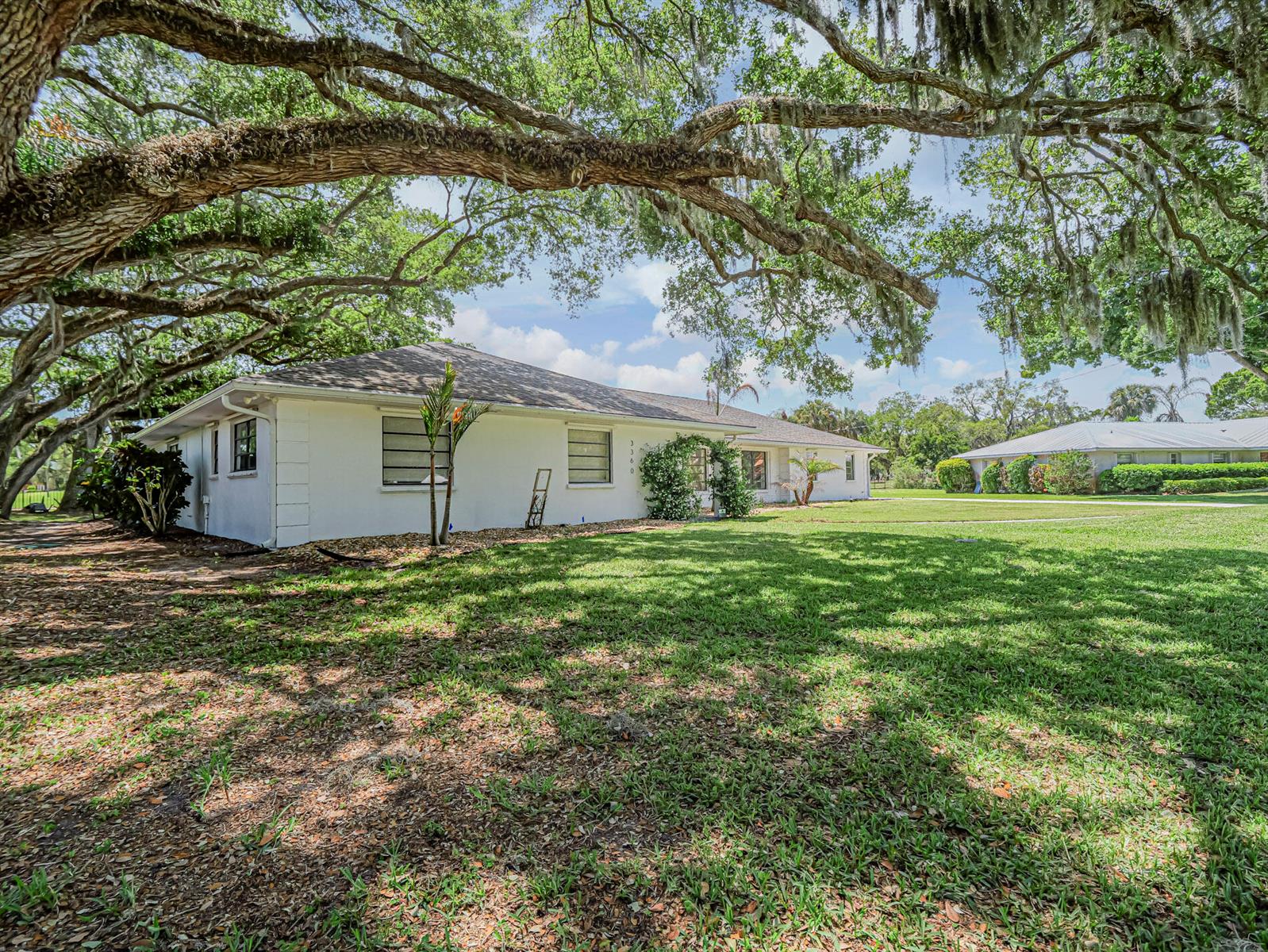 A white single-story house sits on a grassy lawn under large, moss-covered oak trees — 3360 Buckinghammock Trail, Vero Beach FL 32960, VERO BEACH, FL