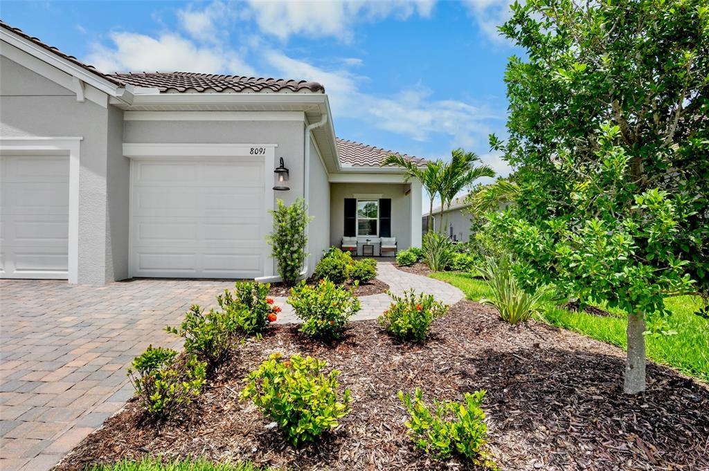 A single-story house exterior features a white garage door, a paved walkway, and landscaping — 8091 ANTHIRIUM LOOP, SARASOTA, FL
