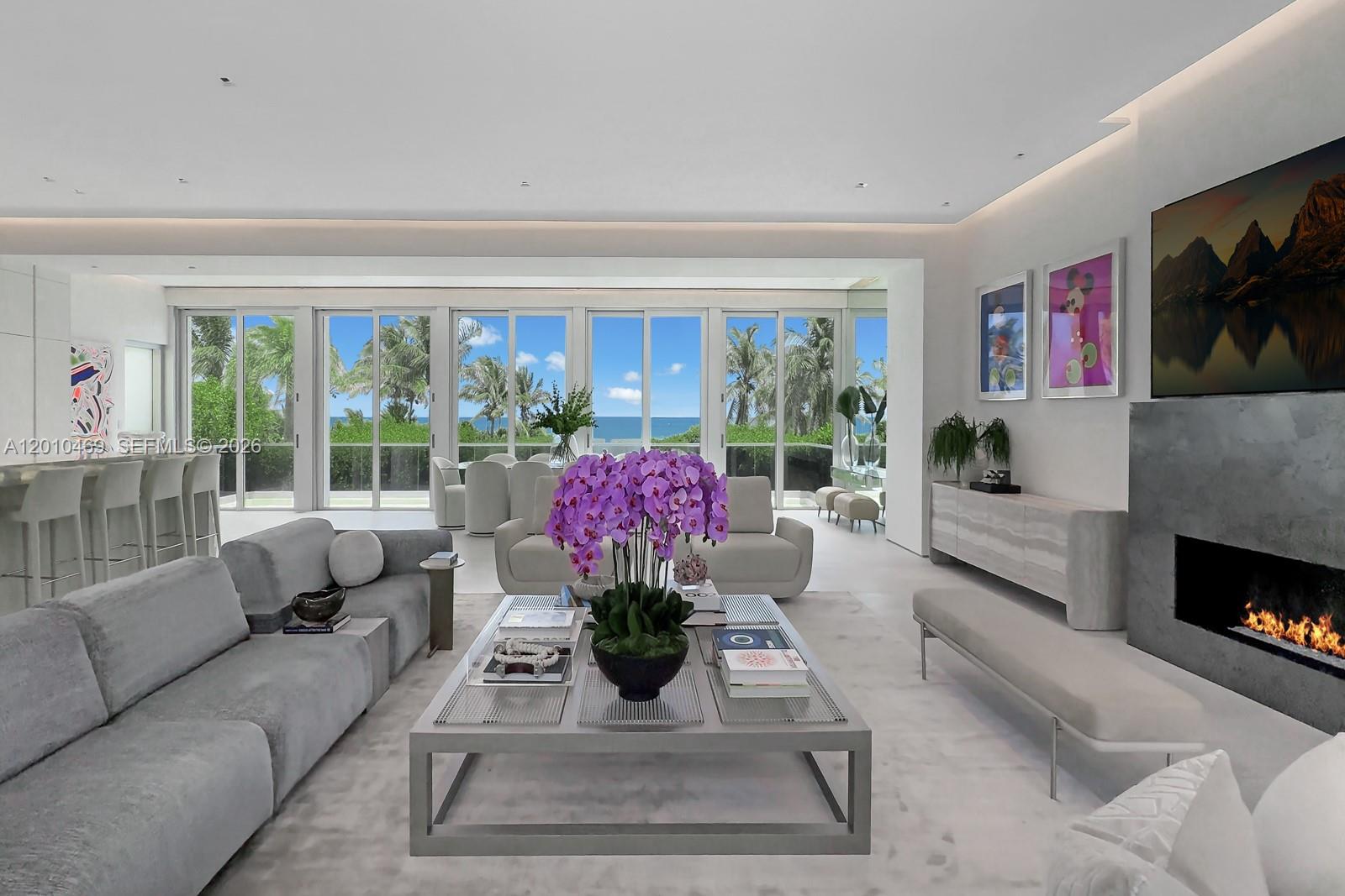 A modern living room features a fireplace, gray sofas, and floor-to-ceiling windows overlooking the ocean — 7815 Atlantic Way, Miami Beach FL 33141, MIAMI BEACH, FL