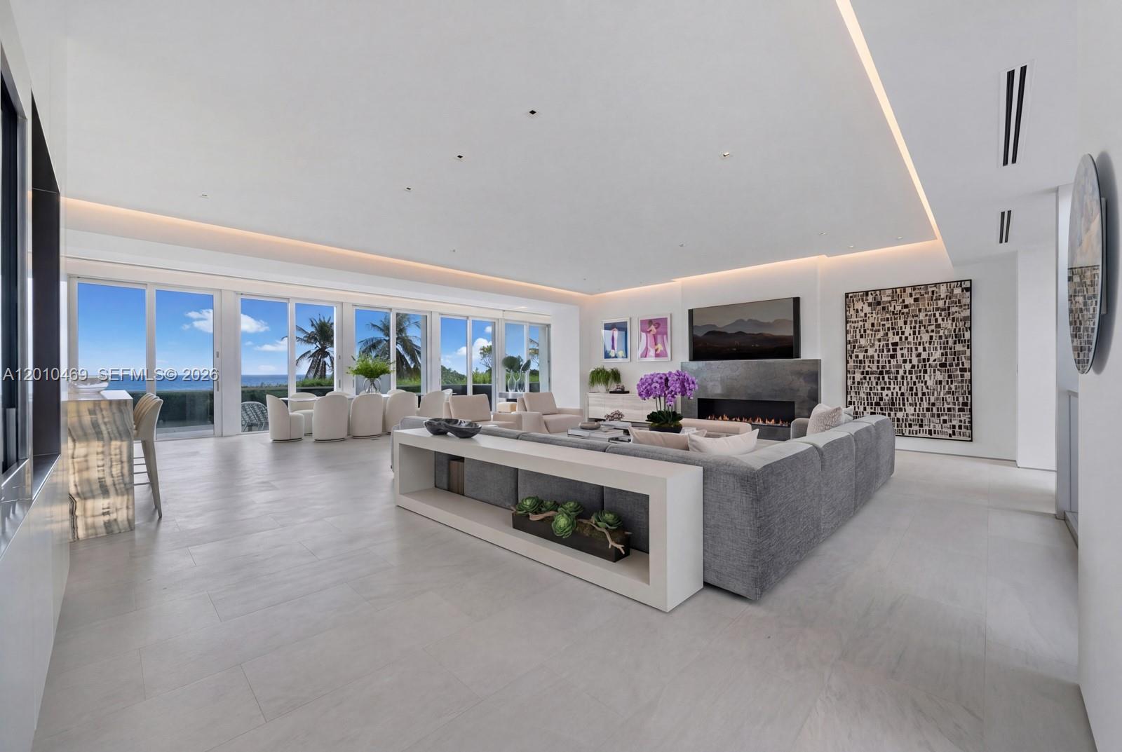 A modern living room features a gray sectional sofa, fireplace, and floor-to-ceiling windows with ocean views — 7815 Atlantic Way, Miami Beach FL 33141, MIAMI BEACH, FL