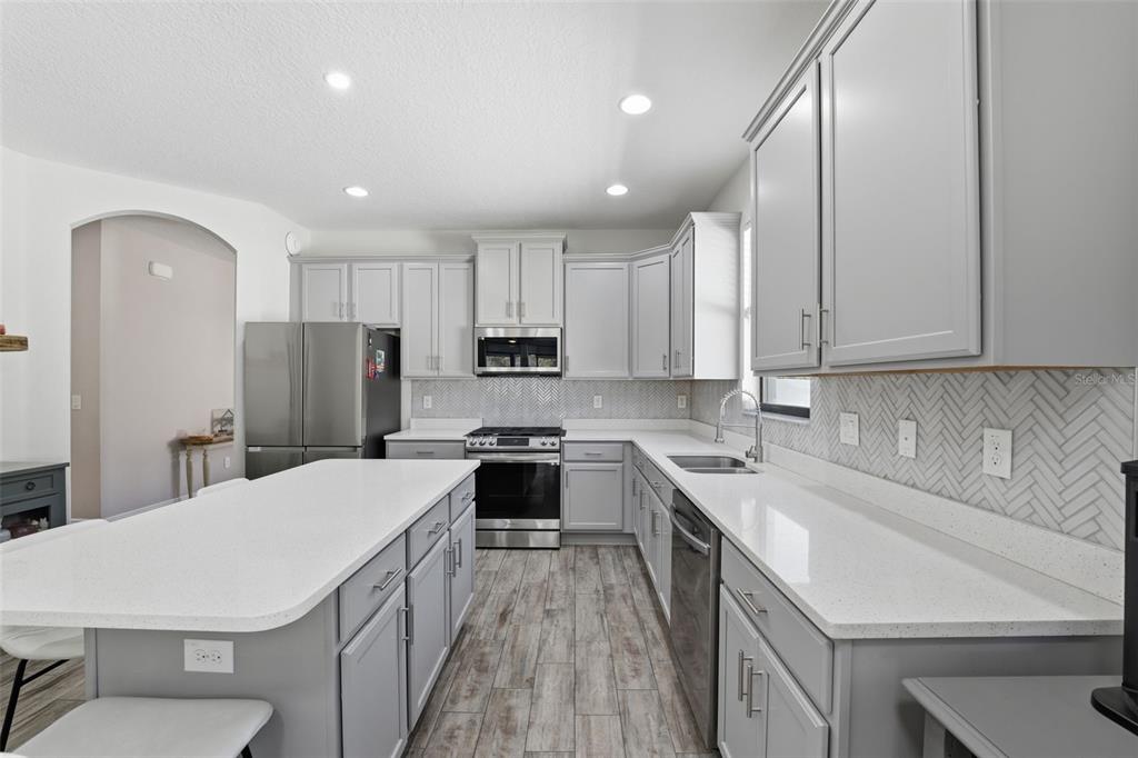 A modern kitchen features gray cabinets, a white island with bar stools, and dining area — 8328 AUBURN RISE COURT, LAND O LAKES, FL