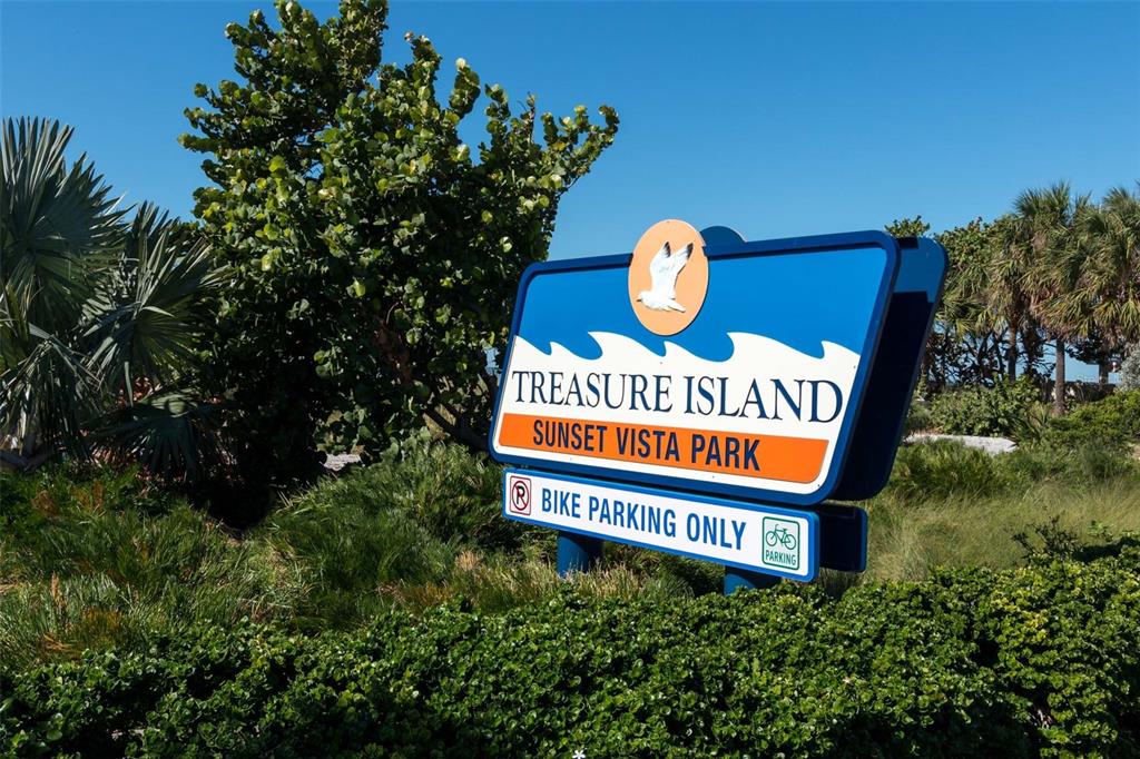 A blue and orange sign for Treasure Island Sunset Vista Park stands amidst green foliage — 9755 HARRELL AVENUE UNIT 102, TREASURE ISLAND, FL