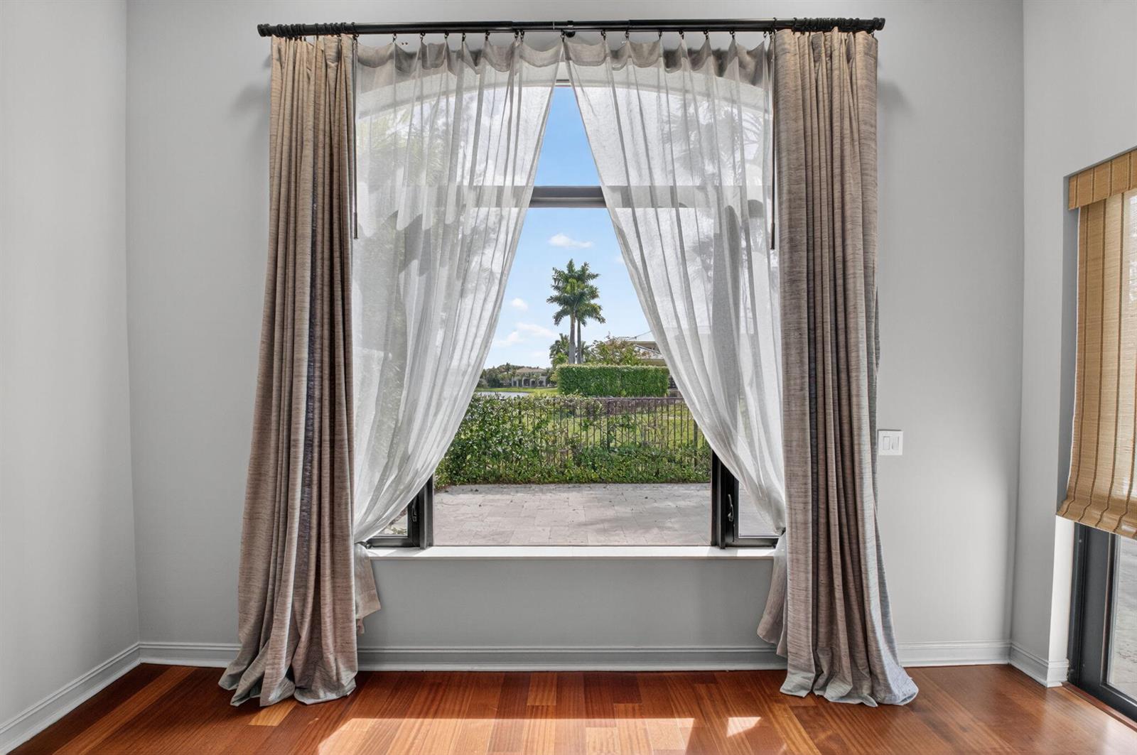 A room with hardwood floors features a large window draped with curtains overlooking a yard — 17921 Villa Club Way, Boca Raton FL 33496, BOCA RATON, FL