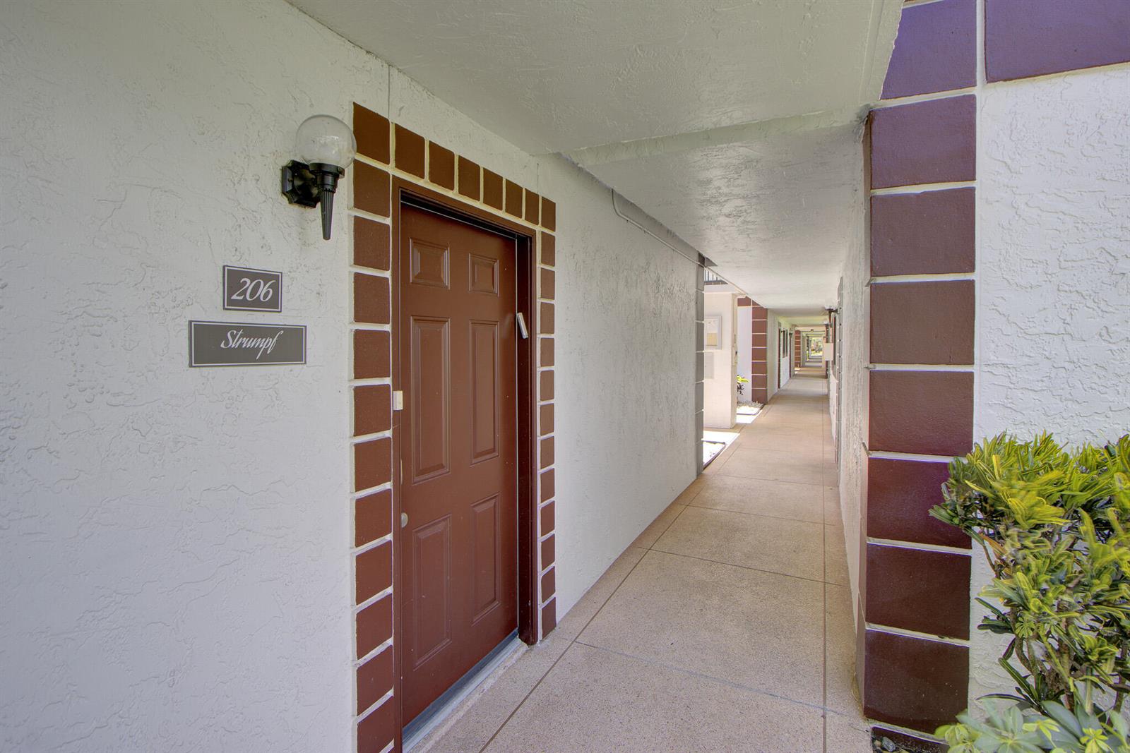 An exterior hallway features a brown door labeled 206 with a nameplate and light fixture — 15451 Pembridge Drive # 206, Delray Beach FL 33484, DELRAY BEACH, FL