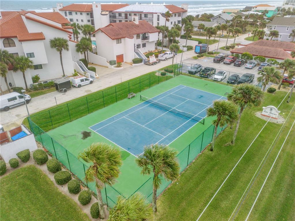 An aerial view shows a fenced tennis court next to a parking lot and buildings — 3001 S ATLANTIC AVENUE UNIT 305, NEW SMYRNA BEACH, FL