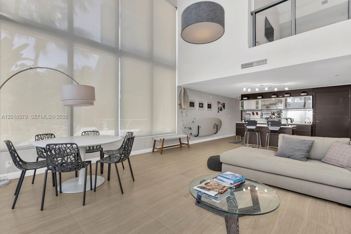 A modern living and dining area features high ceilings, large windows, and light wood flooring — 6000 Collins Ave # 129, Miami Beach FL 33140, MIAMI BEACH, FL