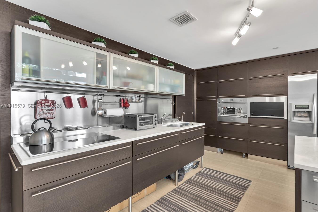 A modern kitchen features dark wood cabinetry, stainless steel appliances, and a white countertop — 6000 Collins Ave # 129, Miami Beach FL 33140, MIAMI BEACH, FL