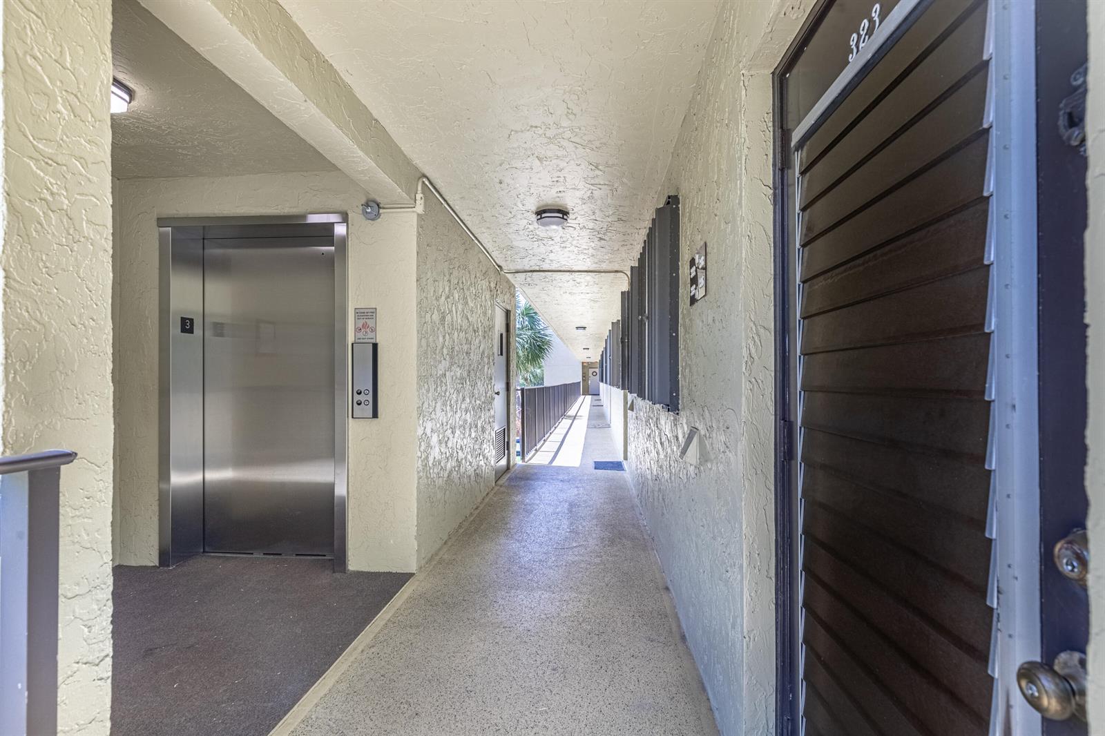 An outdoor apartment hallway features a metal elevator, textured walls, and a dark door — 367 S Federal Highway # C323, Deerfield Beach FL 33441, DEERFIELD BEACH, FL