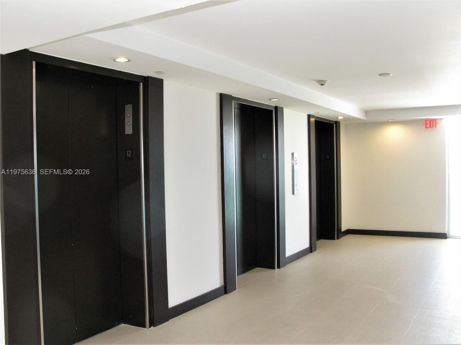A hallway features three black elevator doors with white walls and recessed ceiling lighting — 5701 Collins Ave # 803, Miami Beach FL 33140, MIAMI BEACH, FL