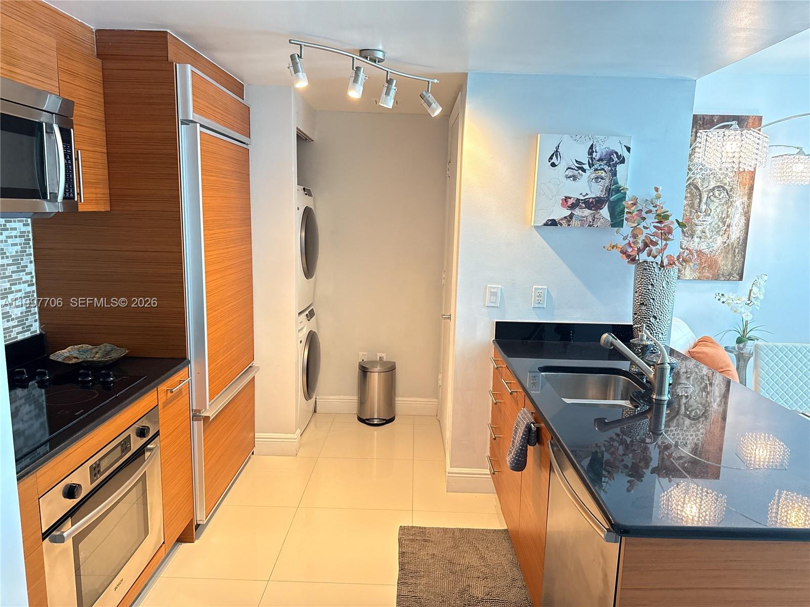 A modern kitchen features wood-paneled cabinetry, black countertops, stainless steel appliances, and a stacked laundry unit — 50 Biscayne Blvd # 1907, Miami FL 33132, MIAMI, FL