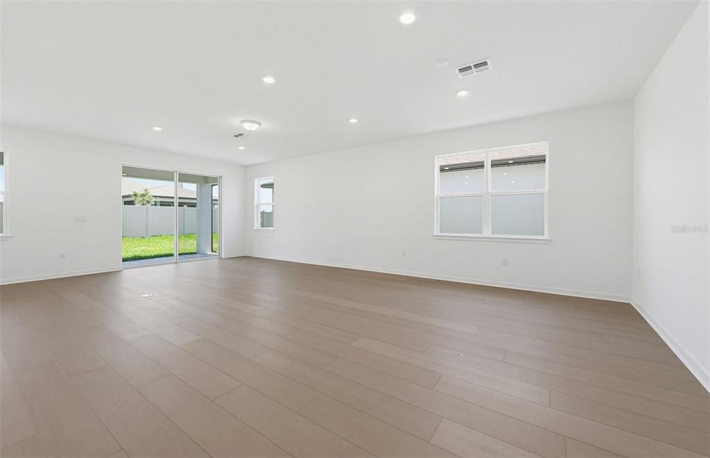 A bright, empty living room features light wood-look flooring, white walls, and a sliding door — 33425 HERDS HALLOW WAY, WESLEY CHAPEL, FL