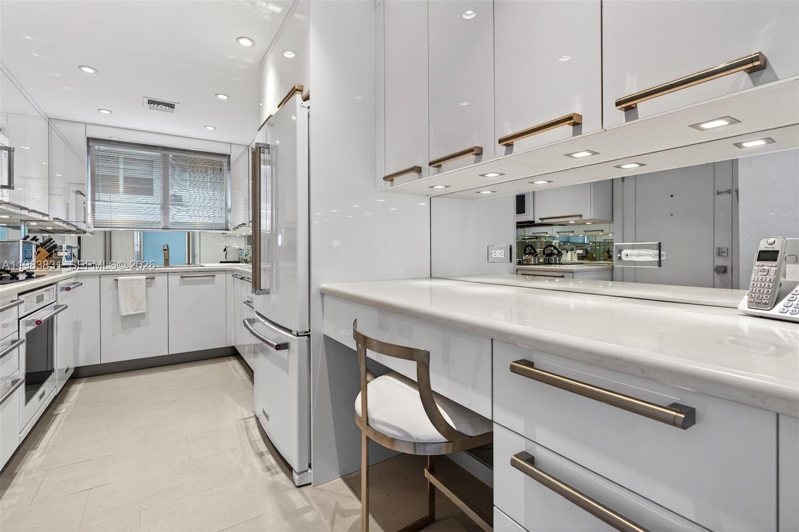 A modern white kitchen features sleek cabinetry, gold hardware, a mirrored backsplash, and a desk area — 10178 Collins Ave # 208, Bal Harbour FL 33154, BAL HARBOUR, FL