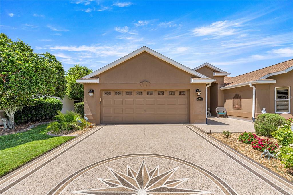 A tan house exterior features a two-car garage and a decorative compass rose driveway design — 8481 SE 168TH KITTREDGE LOOP, THE VILLAGES, FL