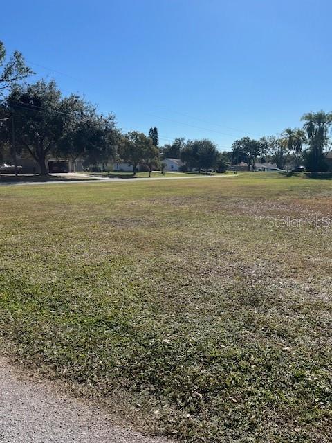 A large, grassy vacant lot is surrounded by trees and residential houses under a clear sky — Lot 589 MARIE DRIVE, ZEPHYRHILLS, FL