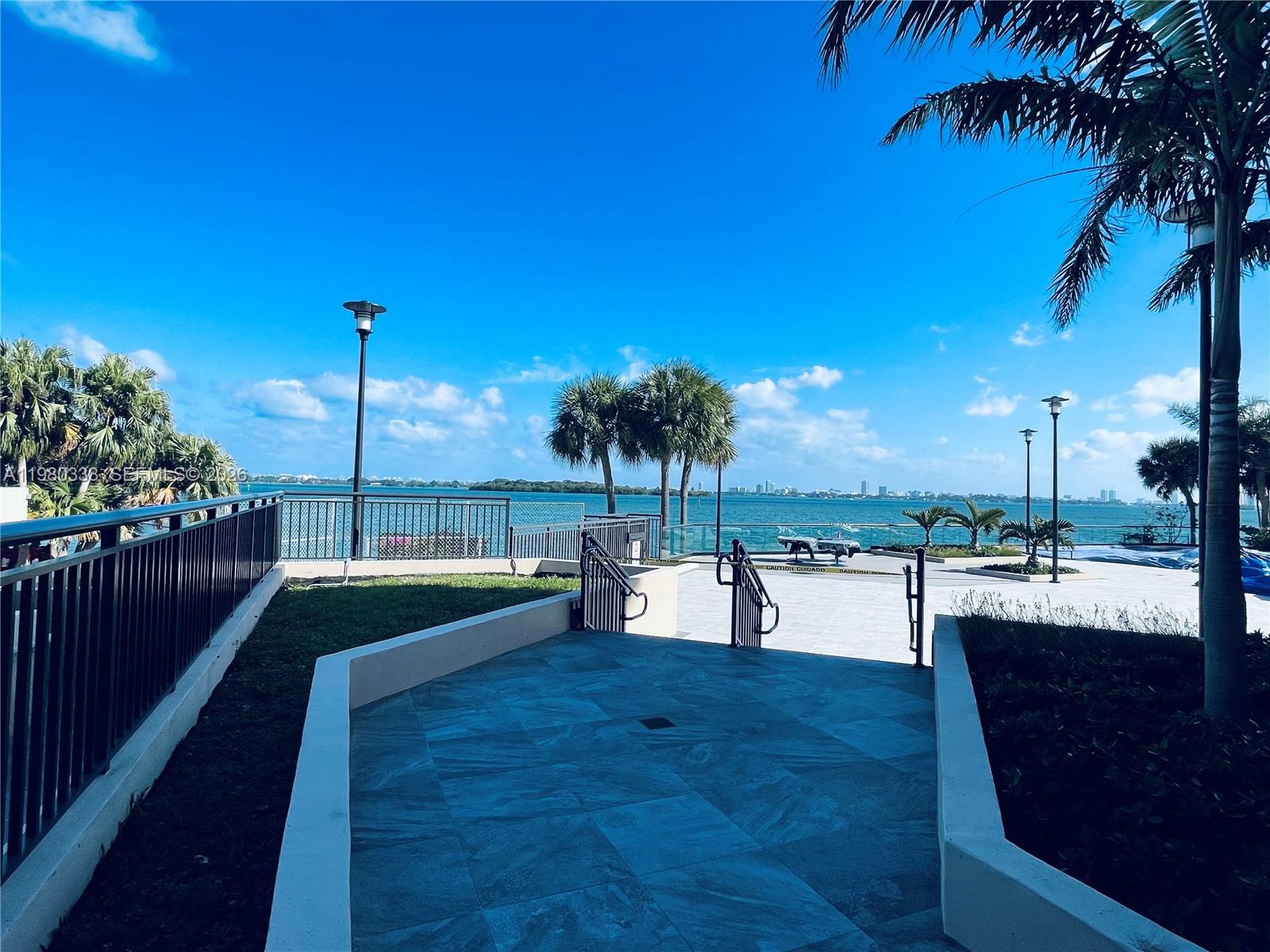 A paved walkway leads to a waterfront terrace with palm trees and a city skyline view — 11111 Biscayne Blvd # 4E, Miami FL 33181, MIAMI, FL