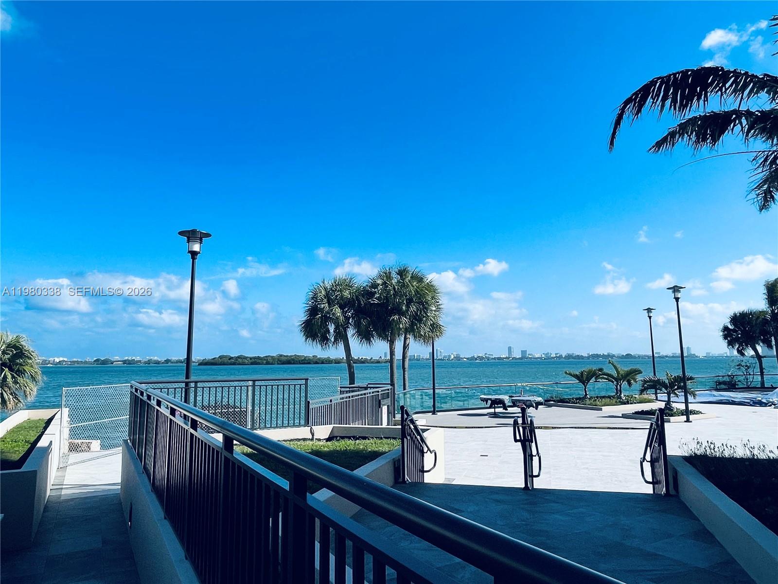 A paved waterfront terrace features metal railings, palm trees, and views of a distant city skyline — 11111 Biscayne Blvd # 4E, Miami FL 33181, MIAMI, FL