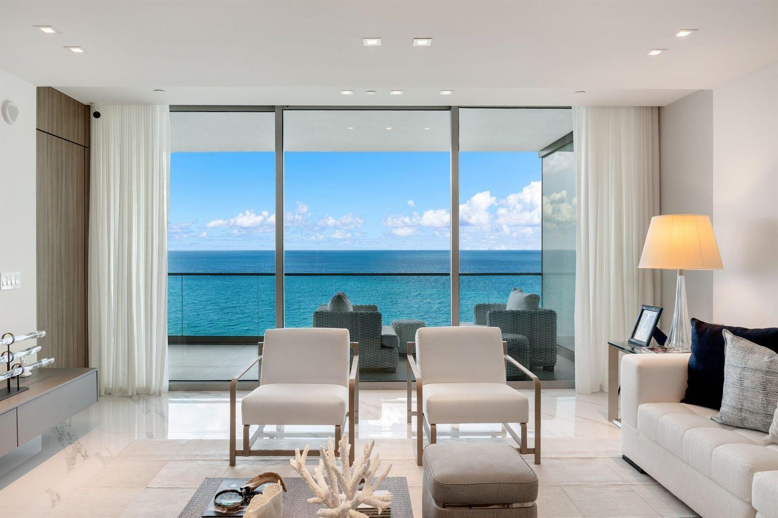 A living room with white chairs and a sofa overlooks the ocean through floor-to-ceiling windows — 10201 Collins Avenue # 1803, Bal Harbour FL 33154, BAL HARBOUR, FL