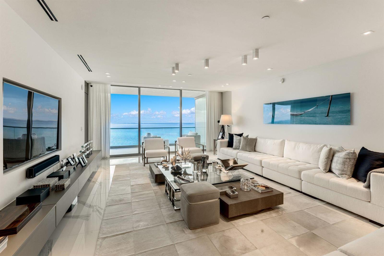 A modern living room features a large white sectional, glass coffee table, and ocean views — 10201 Collins Avenue # 1803, Bal Harbour FL 33154, BAL HARBOUR, FL