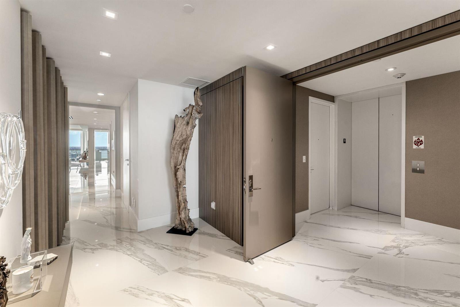 A modern foyer features white marble flooring, a large driftwood sculpture, and a wood-paneled door — 10201 Collins Avenue # 1803, Bal Harbour FL 33154, BAL HARBOUR, FL