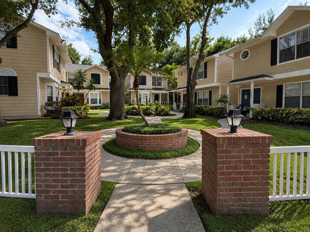 A brick-paved walkway leads through a courtyard between two-story tan townhomes with white fences — 2390 GUN FLINT TRAIL, PALM HARBOR, FL