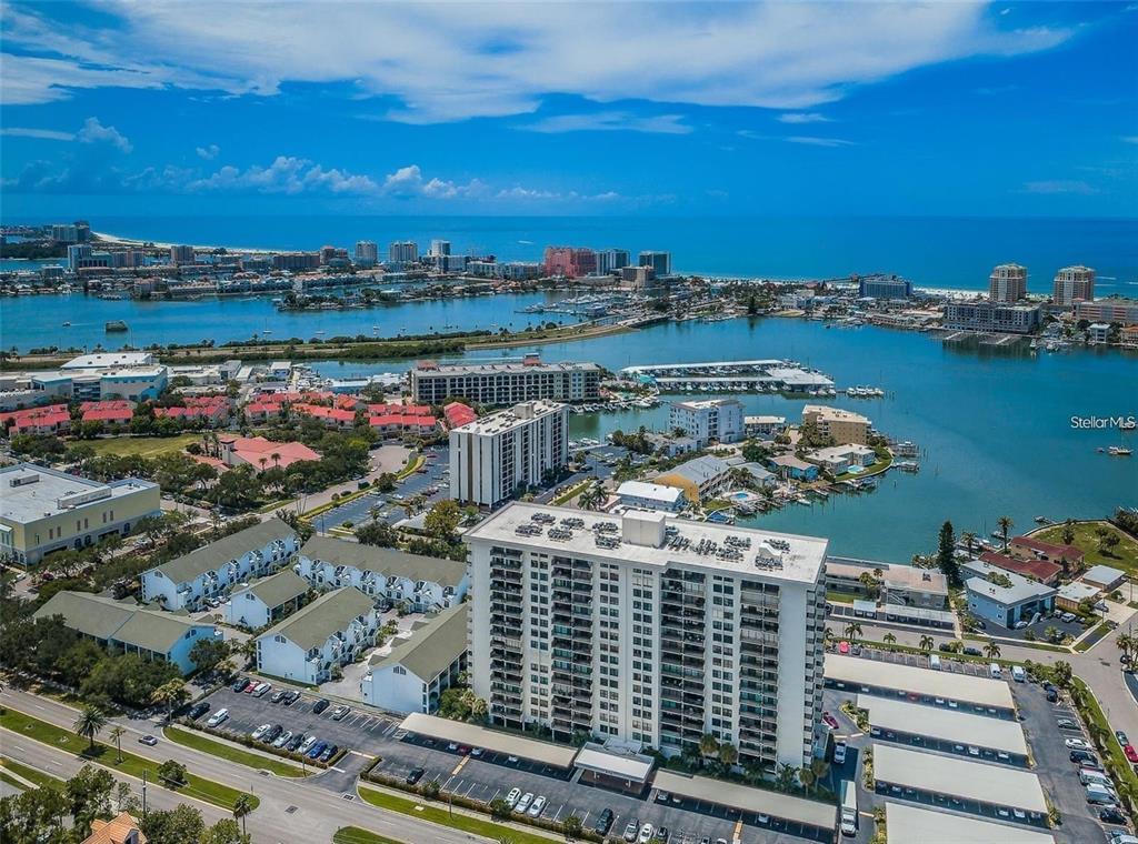 An aerial view shows a high-rise condominium building surrounded by parking lots, residential complexes, and water — 400 ISLAND WAY UNIT 309, CLEARWATER BEACH, FL