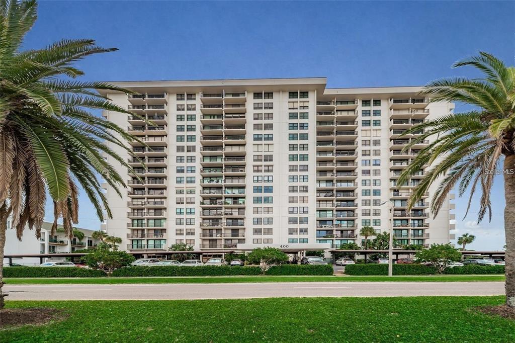 A multi-story residential building with balconies and palm trees stands behind a grassy lawn — 400 ISLAND WAY UNIT 309, CLEARWATER BEACH, FL