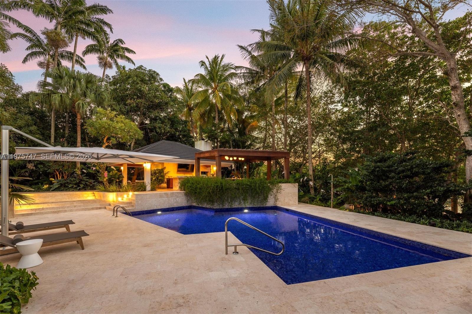 A swimming pool with a stone deck sits in front of a house and pergola — 7150 W Lago Dr, Coral Gables FL 33143, CORAL GABLES, FL
